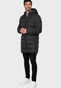 Svart puffjacka med huva, dragkedjestängning och quiltad design. Har sidofickor, bärs över svarta jeans och vita sneakers.