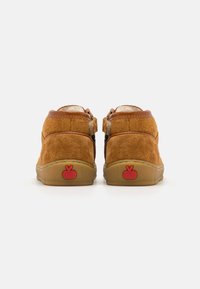 Chaussures en suede tan avec un col en cuir marron, design bas, et un logo rouge rond sur le talon. Texture douce et semelle en caoutchouc.