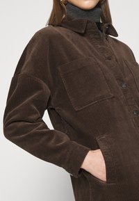 Hnědý kordákový kabát s límečkem, zapínáním na knoflíky, dvěma předními kapsami a ribovanou texturou. Má uvolněný střih a dlouhé rukávy.
