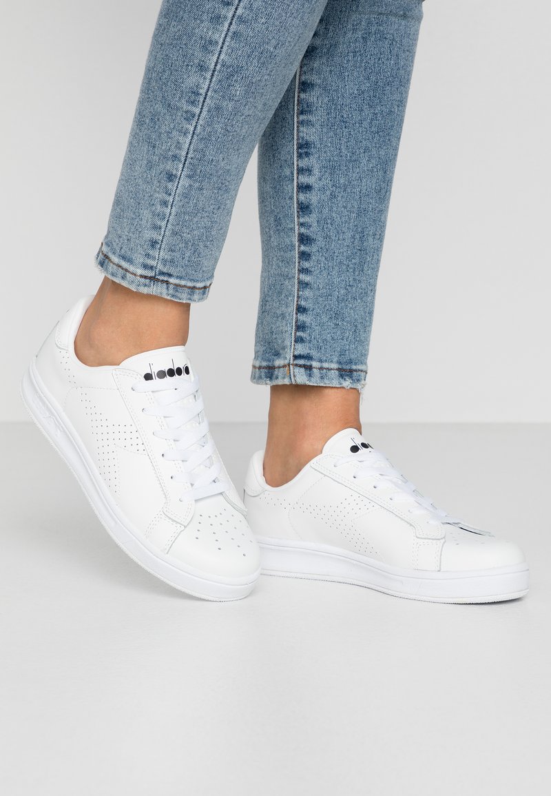 Sneakers bianchi con dettagli perforati sui lati, lacci piatti e una suola bianca texture. Abbinati a jeans blu chiaro con risvolti.