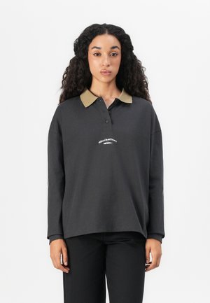 Femme avec de longs cheveux bouclés portant un polo noir ample à manches longues avec un col beige et un pantalon noir, se tenant devant un fond blanc.