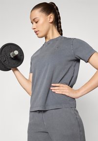 T-shirt en coton gris avec un col rond et des manches courtes, présentant un petit logo. Le modèle tient une plaque de poids noire avec une poignée en argent.