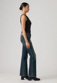 Jean en denim bleu foncé évasé avec une taille mi-haute et des accents usés, associé à un débardeur noir ajusté et des bottes à talons noires.