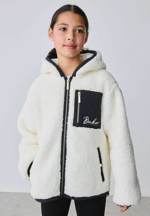 Pige iført en hvid fluffy hættejakke med sort lynlås og brystlomme, står mod en ensartet lys baggrund.