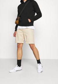 Svart huoodie, vit lager-på-lager skjorta, beiga shorts, svarta strumpor och vita sneakers. Brunt crossover-väska. Canva-bakgrund.
