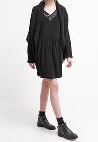 Vestido negro con falda fluida y detalle de encaje en el escote en V, combinado con un cárdigan negro texturizado; acompañado de botas de tobillo negras con tachuelas.