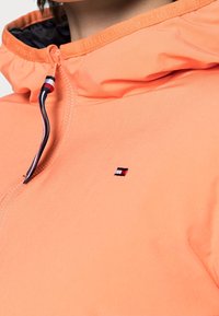 Primo piano di una persona che indossa una giacca a cappuccio pesco con zip e un piccolo logo rosso, bianco e nero sul petto.