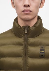 Jeune homme portant une doudoune vert olive fermée par une fermeture éclair avec un col montant, affichant une étiquette de la marque Blauer sur la poitrine.