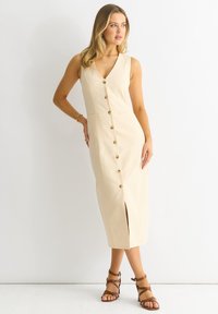 Beige, mouwloze midi-jurk met een V-hals, knoopsluiting aan de voorkant, een zijsplit en zakken. Gestyled met bruine sandaaltjes met bandjes.