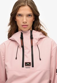 Lys rosa pullover med høj krave, sort lynlås og justerbare snore. Har et logopatch og sorte accenter. Glat tekstur.