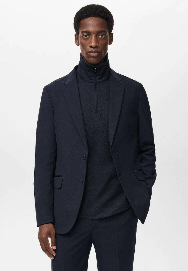 Giacca da abito blu navy con rever a taglio e due bottoni, abbinata a un top in maglia con colletto alto. Tessuto dalla superficie liscia, vestibilità sartoriale.