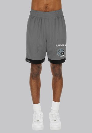 Mann trägt graue Mesh-Sportshorts mit schwarzem Besatz, weiße Socken, weiße Sneakers und "Raiders" Text mit Football-Helm-Logo auf den Shorts.