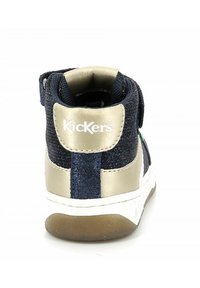 Baskets montantes noires et dorées avec un tissu texturé, des accents métalliques dorés, une bande Velcro et une semelle en caoutchouc blanche. Présente le logo Kickers sur le talon.