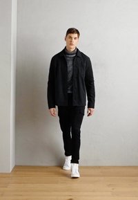 Svart knappjakke over en teksturert grå genser, paret med svarte bukser og hvite sneakers, stående på et tregulv mot en grå vegg.