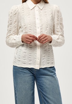 Femme portant un chemisier blanc texturé à boutons avec col et manches retroussées, associé à un jean bleu, mains jointes devant.