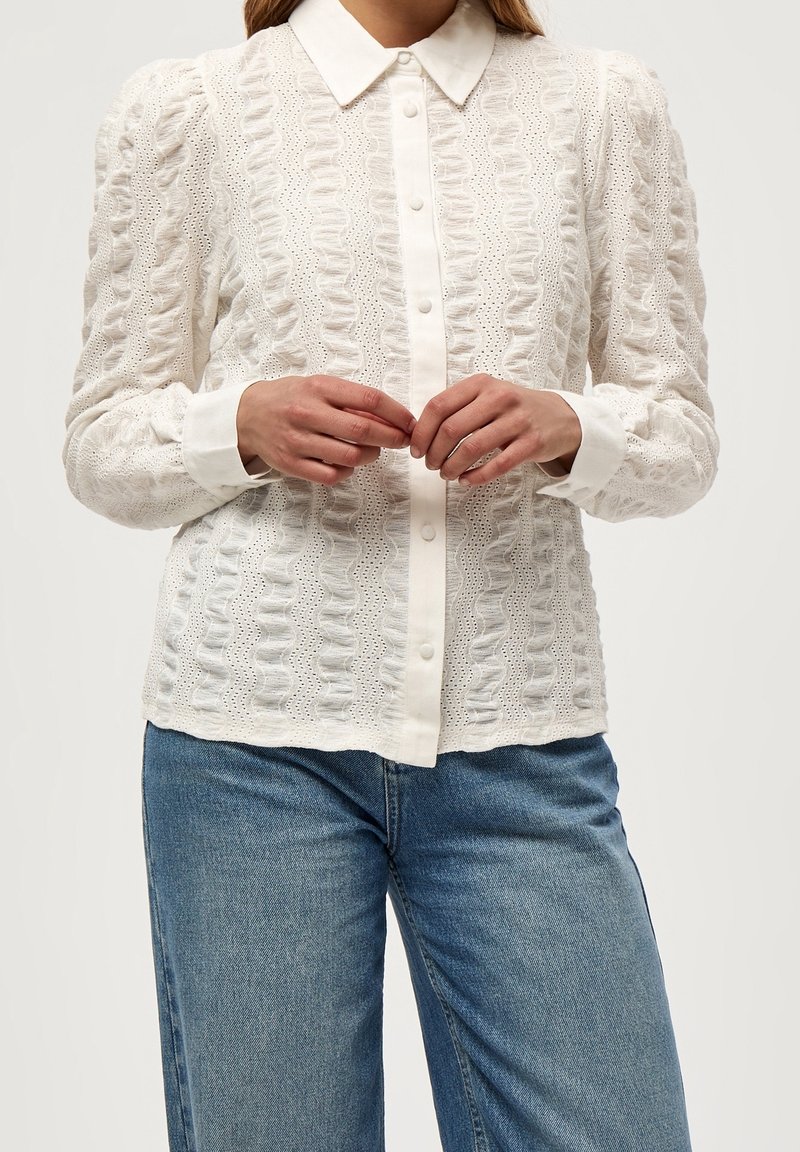 Femme portant un chemisier blanc texturé à boutons avec col et manches retroussées, associé à un jean bleu, mains jointes devant.