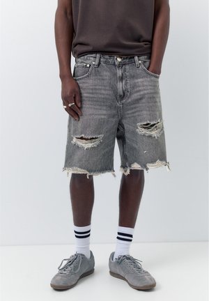 Férfi szürke, szakadt farmer rövidnadrágban, sötét pólóban, fehér fekete csíkos zokniban, szürke sportcipővel és ezüst gyűrűkkel a bal kézen.