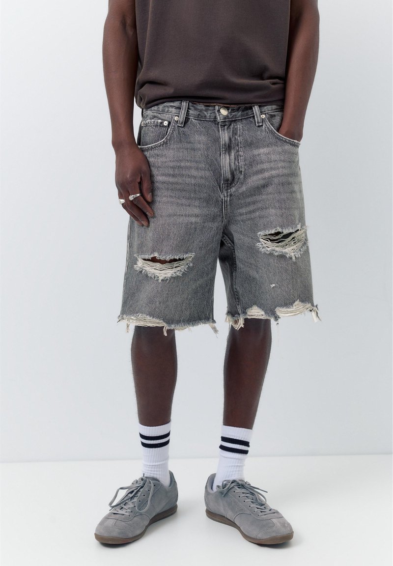 Homme portant un short en jean gris déchiré, un t-shirt foncé, des chaussettes blanches à rayures noires, des baskets grises et des bagues argentées à la main gauche.