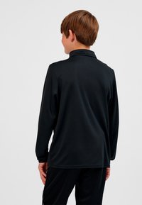 Junge mit hellbraunen Haaren, der ein schwarzes langärmeliges Hemd und schwarze Hosen trägt, mit dem Rücken zu uns vor einem schlichten hellen Hintergrund.