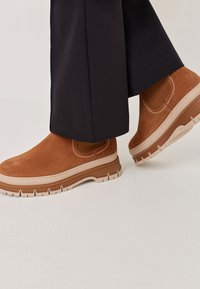 Hnědé semišové kotníkové boty s béžovými chunky podrážkami, nošené s černými širokými kalhotami na světlejší podlaze.