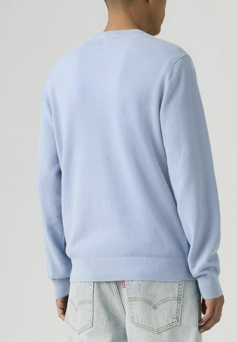 Hombre vistiendo un suéter de manga larga de color azul claro con textura y jeans de mezclilla en tono claro, mostrado de espaldas sobre un fondo liso.