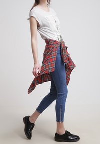 Ležérní outfit sestává z bílého trika s krátkým rukávem, modrých skinny džínů se zipy na kotnících a uvázané červené kostkované flanelové košile. Černé šněrovací boty.