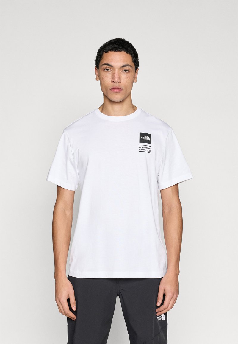 T-shirt en coton blanc à manches courtes, col rond, avec un patch logo noir sur le côté gauche de la poitrine. Texture lisse, coupe décontractée.