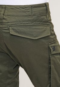 Pantalon cargo vert olive avec deux poches arrière à rabat et une poche latérale. Le tissu est robuste avec une texture lisse et des coutures renforcées.