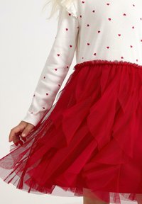 Weißes Langarmoberteil mit roten Herzmustern, kombiniert mit einem roten, geschichteten Tüllrock mit Rüschensaum und elastischem Bund.
