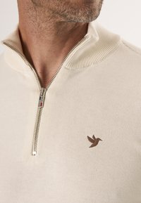 Beiger Strickpullover mit hohem Kragen und silbernem Reißverschluss, mit einem kleinen braunen Vogel-Logo auf der Brust. Weiche Textur und taillierte Passform.