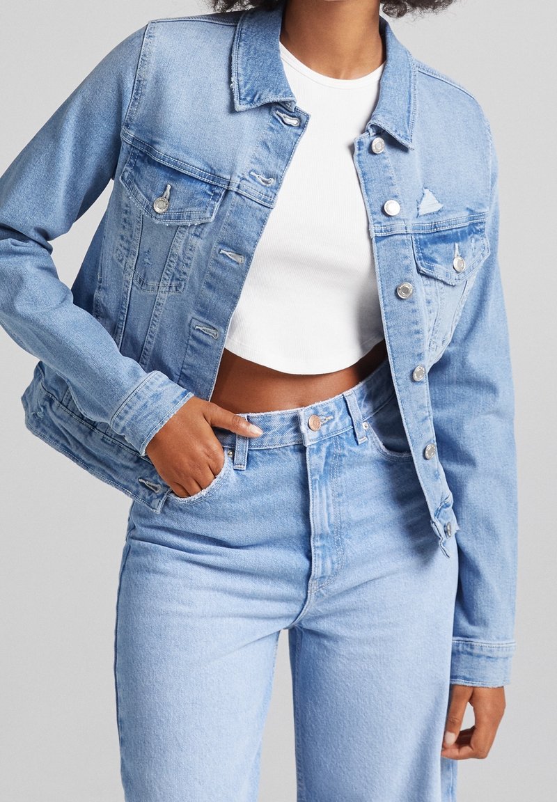 Giacca di jeans azzurro chiaro con colletto classico, tasche frontali e dettagli strappati. Indossata con un top bianco corto e jeans in denim coordinati.