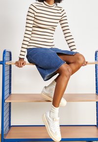 Gestreiftes Oberteil mit Stehkragen in Creme mit gelben und blauen Streifen, kombiniert mit einem knielangen Jeansrock und weißen Sneakern mit beigen Akzenten.