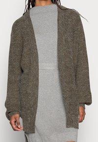 Hnědý pletený kardigan s otevřeným předním dílem, oversized rukávy a texturovaným povrchem. Nesený přes světle šedé žebrované šaty, vytváří vrstvený vzhled.