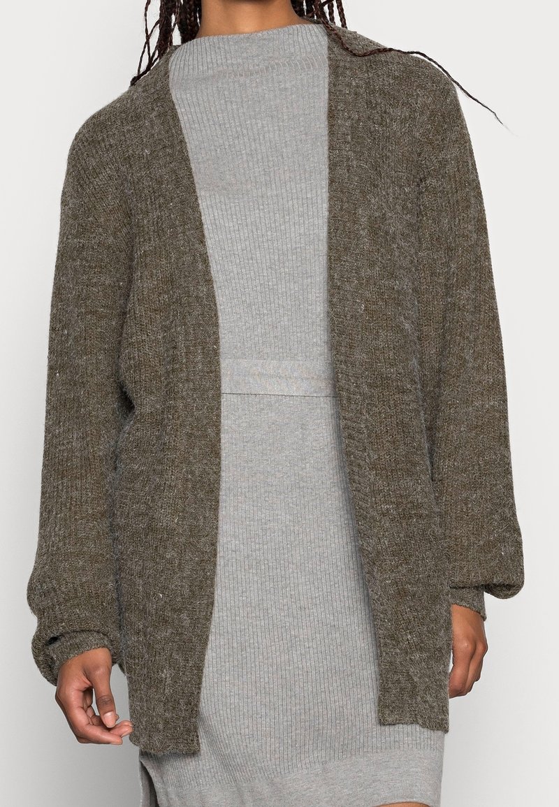 Hnědý pletený kardigan s otevřeným předním dílem, oversized rukávy a texturovaným povrchem. Nesený přes světle šedé žebrované šaty, vytváří vrstvený vzhled.