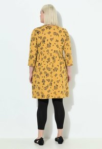 Donna con capelli biondi lisci indossa un vestito giallo con motivo paisley e maniche a tre quarti, leggings neri e scarpe ballerine nere, in piedi di fronte a un muro.