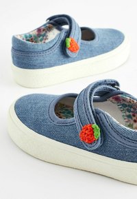 Chaussures en denim bleu pour enfants avec semelles blanches, une sangle à boucle et un accent en fraise crochetée sur le côté. Doublure florale visible.