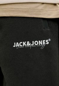 Zwarte katoenen joggers met een logo in witte tekst, "JACK&JONES," nabij de bovenkant, met daaronder een kleinere slogan. De stof lijkt zacht.