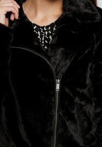 Woman wearing a black faux fur jacket with a diagonal zipper over a black and white patterned top, holding the collar with one hand.