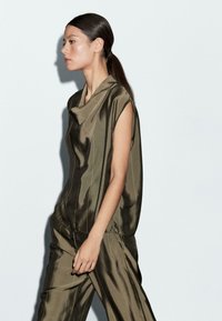 Vrouw met een donkere paardenstaart, gekleed in een losse, glanzende olijfgroene mouwloze top en bijpassende wijde broek, die loopt tegen een effen lichte achtergrond.