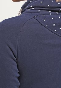 Mörkblå sweatshirt med en mönstrad huva med små vita motiv, slät textur och en etikett på baksidan nära halslinningen.