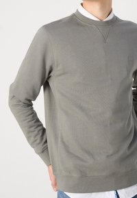 Man som bär en ljus olivgrön BOSS-tröja över vit skjorta med blå jeans, hand vilande på höften, kortad vid halsen och höften.