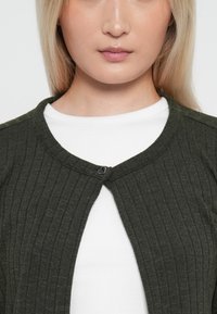 Mørkegrønn ribbestrikket cardigan med rund hals og en dekorativ metallkrok, båret over en hvit topp med teksturert overflate.