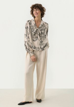Blouse in beige with black paisley pattern, featuring a tie neckline and long sleeves. Paired with lightweight beige trousers and black flats.