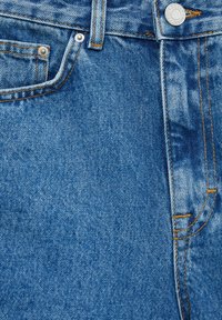 Gros plan sur un jean en denim bleu montrant la poche avant, la boucle de ceinture, les rivets métalliques, les surpiqûres et le bouton argenté avec texte sur la ceinture.