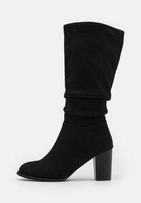 Bottine noire en daim avec une tige souple, un bout pointu et un talon haut épais. Texture lisse avec une finition douce.