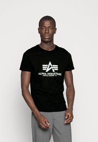 Black cotton t-shirt with white printed logo design featuring "ALPHA INDUSTRIES" and geometric accents. Round neckline, short sleeves.