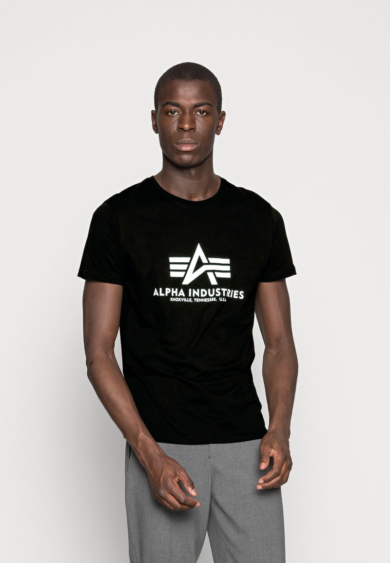 Black cotton t-shirt with white printed logo design featuring "ALPHA INDUSTRIES" and geometric accents. Round neckline, short sleeves.