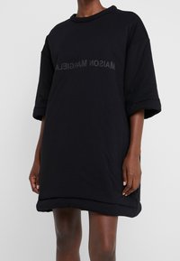 Czarna oversize sukienka z miękkiej bawełny, z rolowanym dekoltem i krótkimi rękawami. Z przodu znajduje się napis "MAISON MARGIELA" w kolorze szarym.