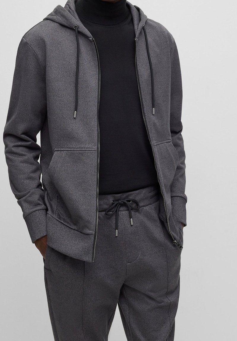 Homme portant un sweat à capuche gris foncé zippé et un pantalon de survêtement assorti avec cordon, au-dessus d'un col roulé noir, mains dans les poches, debout devant un fond uni.