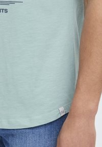 Lys teal kortermet t-skjorte med rund hals, med en subtil tekstur og en merkelapp på kanten, i kombinasjon med blå denimjeans.